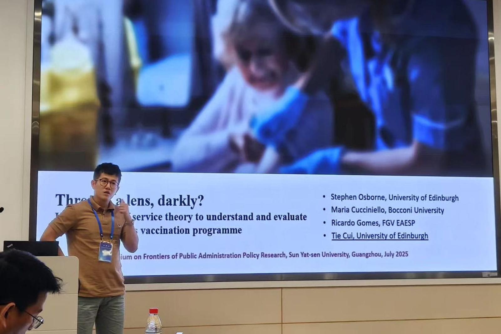 Tie Cui stands presenting at a conference with a large projection screen in the background displaying his presentation slides.